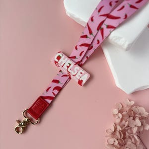 May include: A pink lanyard with a red chili pepper pattern. The lanyard has a gold-colored clasp and a red leather accent. The name "CHELSEA REGISTERED NURSE" is printed on a pink rectangular tag.