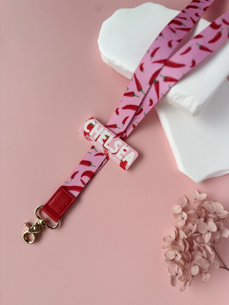 May include: A pink lanyard with a red chili pepper pattern. The lanyard has a gold-colored clasp and a red leather accent. The name "CHELSEA REGISTERED NURSE" is printed on a pink rectangular tag.