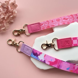 May include: Three colorful lanyards with gold-colored clasps and leather accents. One lanyard is pink with a seashell pattern, another is checkered, and the third has a watercolor floral design. The lanyards are displayed on a pink surface.