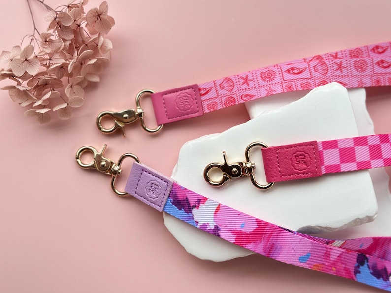May include: Three colorful lanyards with gold-colored clasps and leather accents. One lanyard is pink with a seashell pattern, another is checkered, and the third has a watercolor floral design. The lanyards are displayed on a pink surface.