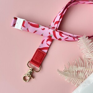 May include: Pink lanyard with a repeating pattern of red chili peppers. The lanyard has a red leatherette accent with a gold-colored clasp. The lanyard is designed to hold keys or an ID badge.