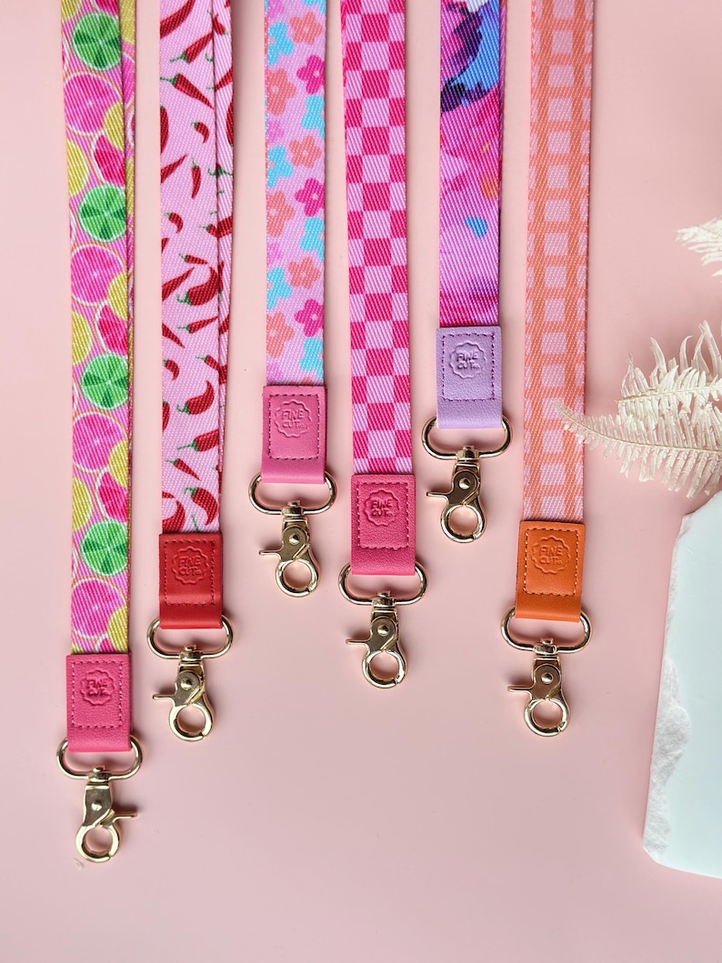 May include: A collection of colorful lanyards with various patterns, including citrus slices, chili peppers, floral designs, and checkered patterns. Each lanyard has a different color leather tab and a gold-colored metal clasp. The lanyards are displayed against a pink background.