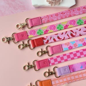 May include: A collection of colorful lanyards with gold-toned clasps. The lanyards feature various patterns, including citrus fruits, chili peppers, and checkered designs. Each lanyard has a leather accent in a coordinating color, such as pink, red, and orange.