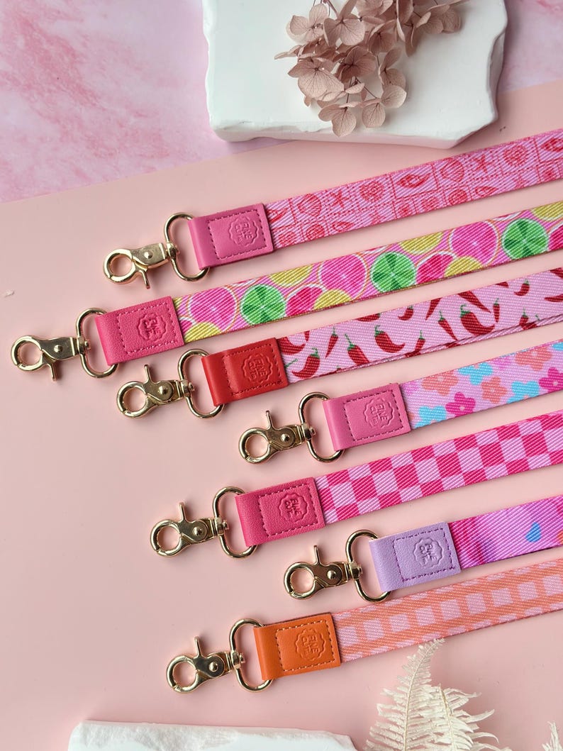 May include: A collection of colorful lanyards with gold-toned clasps. The lanyards feature various patterns, including citrus fruits, chili peppers, and checkered designs. Each lanyard has a leather accent in a coordinating color, such as pink, red, and orange.