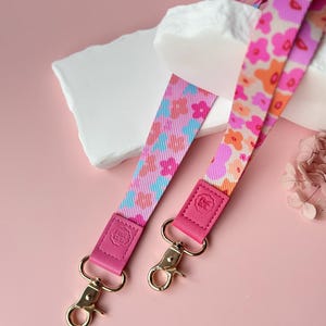 May include: Two floral patterned lanyards with pink, orange, and blue designs. The lanyards have pink leather accents and gold-colored metal clasps. The background is a soft pink.