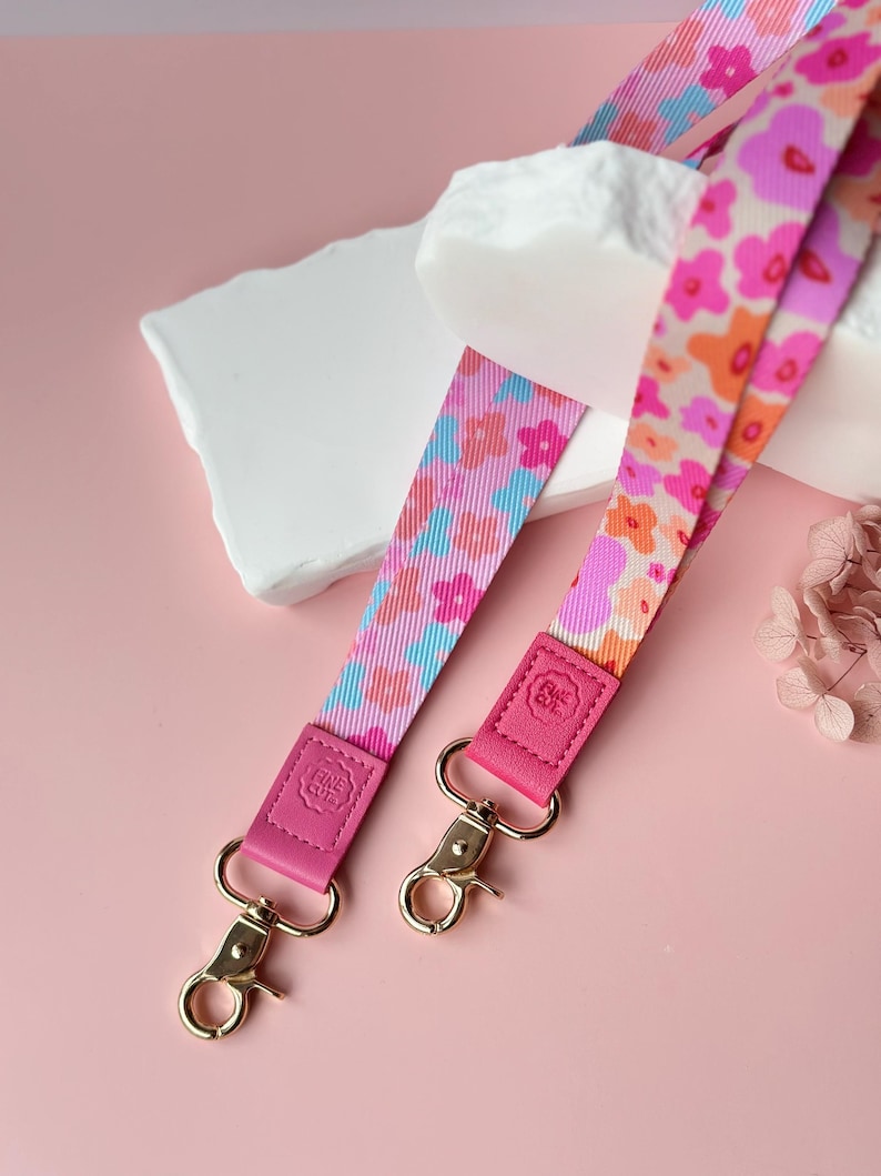 May include: Two floral patterned lanyards with pink, orange, and blue designs. The lanyards have pink leather accents and gold-colored metal clasps. The background is a soft pink.