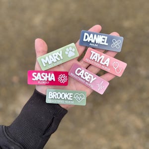May include: A selection of rectangular name badges in various colours, held in a hand. Each badge displays a name and profession, such as "Mary, Vet Nurse" and "Sasha, Florist." The badges have white text and include icons like a paw print and a stethoscope.