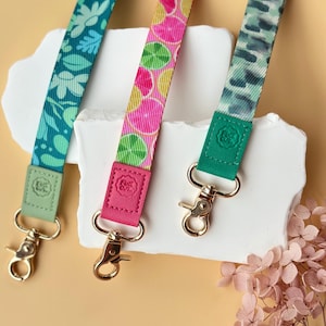 May include: Three colorful lanyards with gold-colored clasps. One is teal with a floral pattern, another is pink with citrus fruit slices, and the third is green with a watercolor-like design. Each lanyard has a matching leather-like accent.