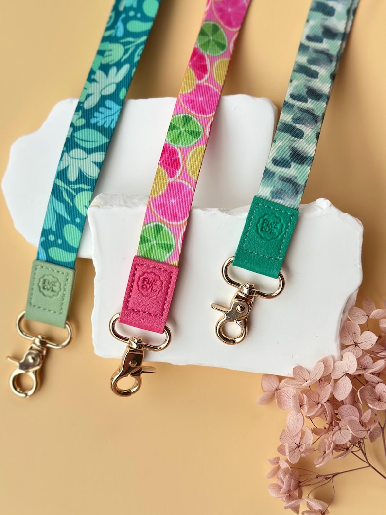 May include: Three colorful lanyards with gold-colored clasps. One is teal with a floral pattern, another is pink with citrus fruit slices, and the third is green with a watercolor-like design. Each lanyard has a matching leather-like accent.