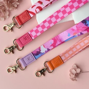 May include: Four colorful lanyards with gold-colored clasps and leather accents. The lanyards feature various patterns, including chili peppers, checkered squares, and abstract designs. The leather accents are in shades of red, pink, purple, and orange.