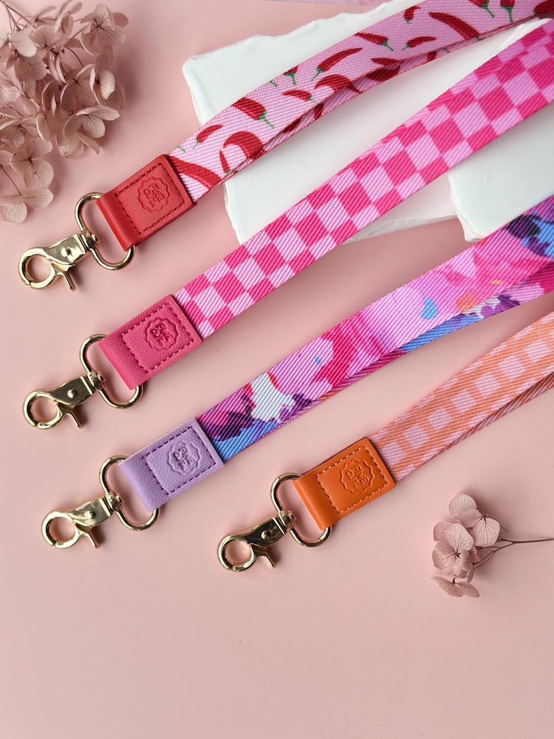 May include: Four colorful lanyards with gold-colored clasps and leather accents. The lanyards feature various patterns, including chili peppers, checkered squares, and abstract designs. The leather accents are in shades of red, pink, purple, and orange.