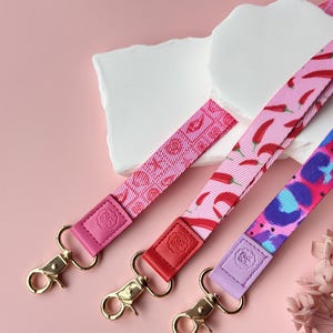 May include: Three colorful lanyards with gold-colored clasps. The lanyards feature different patterns: pink with lips, pink with red chili peppers, and blue and purple leopard print. Each lanyard has a leather accent in pink, red, and purple.