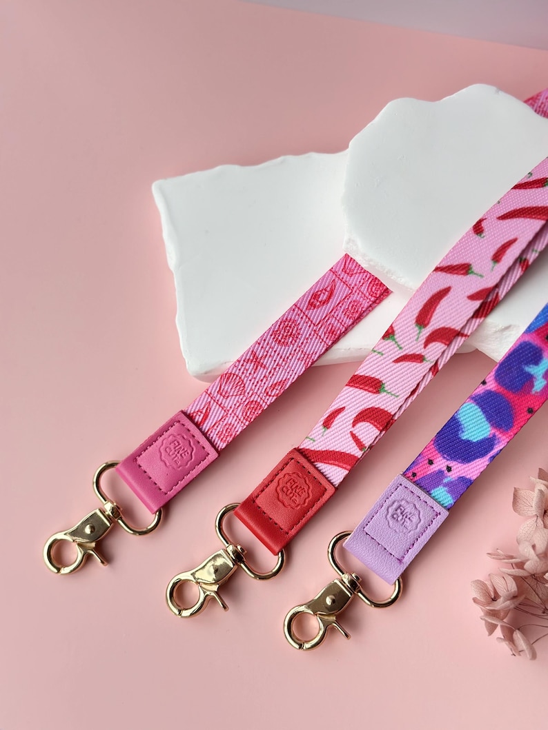May include: Three colorful lanyards with gold-colored clasps. The lanyards feature different patterns: pink with lips, pink with red chili peppers, and blue and purple leopard print. Each lanyard has a leather accent in pink, red, and purple.