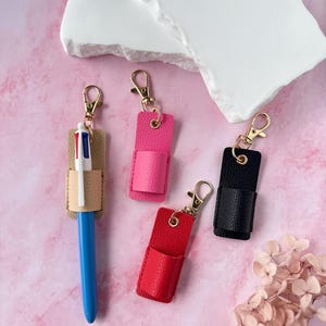 May include: Leather pen holders in pink, red, black, and beige, each with a gold-coloured clasp. A blue pen is inserted into the beige holder. The holders are displayed on a pink surface with a white decorative element.