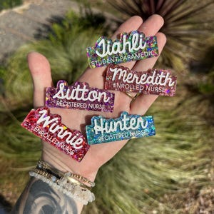 May include: Six colourful glitter resin name tags with white text. The names are Jiahli, Sutton, Meredith, Winnie, Hunter. The text below the names indicates their profession, such as "Student Paramedic", "Registered Nurse", and "Enrolled Nurse".