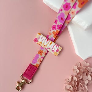 May include: A floral lanyard with the name "Vivian" printed on a rectangular tag. The lanyard features a pattern of pink and orange flowers on a cream background. The lanyard has a pink leather end with a gold-colored clasp.