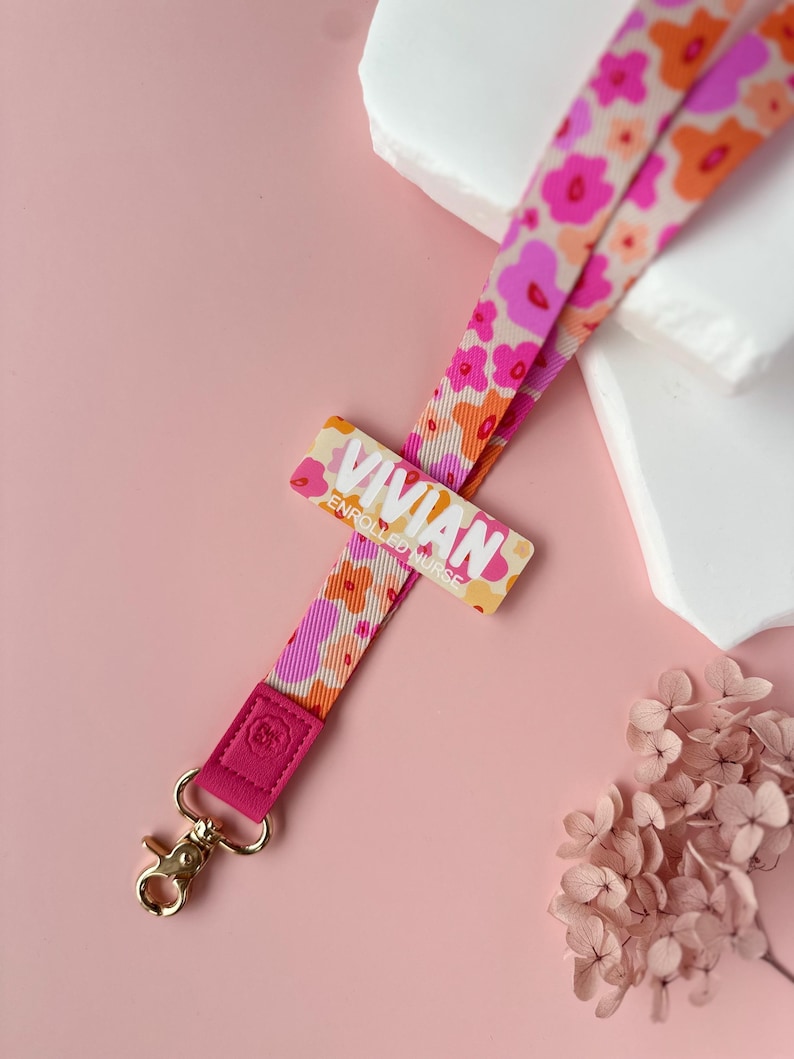 May include: A floral lanyard with the name "Vivian" printed on a rectangular tag. The lanyard features a pattern of pink and orange flowers on a cream background. The lanyard has a pink leather end with a gold-colored clasp.