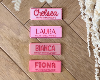 May include: Four pink name badges with white text on a wooden surface. The badges read "Chelsea, Nurse/Midwife", "Laura, Registered Nurse", "Bianca, Speech Pathologist", and "Fiona, Registered Nurse". Decorative white flowers are in the background.
