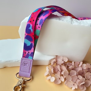 May include: A colorful lanyard with a vibrant pattern of pink, blue, and purple shapes. The lanyard has a lavender leather tab with a gold-colored clasp. Dried pink flowers are in the background.