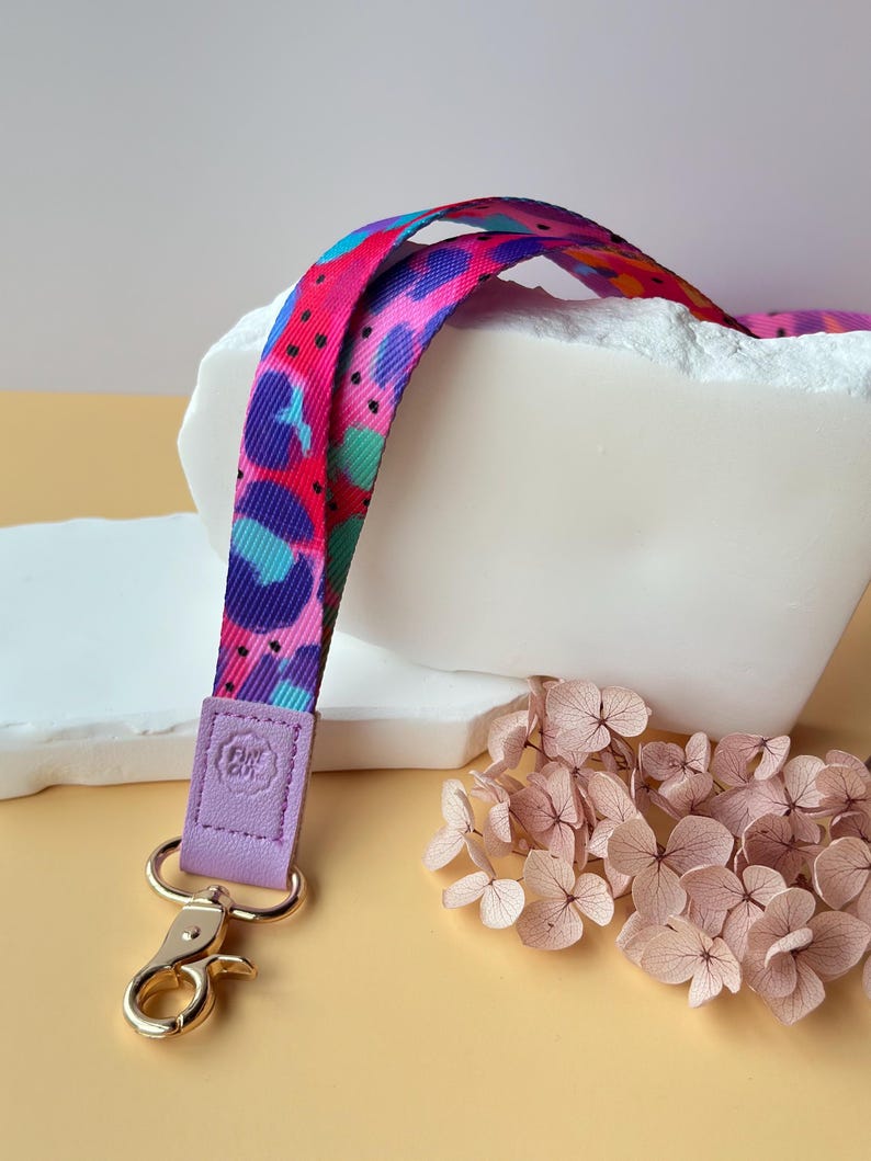 May include: A colorful lanyard with a vibrant pattern of pink, blue, and purple shapes. The lanyard has a lavender leather tab with a gold-colored clasp. Dried pink flowers are in the background.