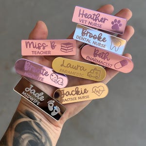 Puede incluir: Una colección de diez etiquetas de nombre de acrílico personalizadas con varios títulos de trabajo e iconos. Las etiquetas son de color rosa, dorado y plateado con letras blancas. Los iconos incluyen un libro, un diente, una píldora, un bebé, un corazón y una huella de pata.