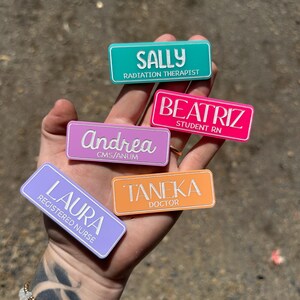 May include: Five colourful name tags with white text. The name tags are made of plastic and have rounded corners. The names on the tags are Sally, Radiation Therapist, Andrea, CMS/ANUM, Beatriz, Student RN, Laura, Registered Nurse, and Tanek, Doctor.