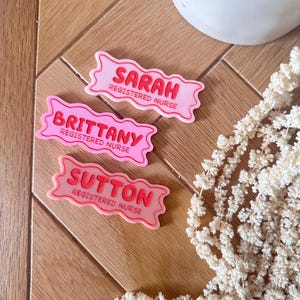 May include: Three pink name tags with white lettering. The name tags are shaped like a banner and have the text "Sarah Registered Nurse", "Brittany Registered Nurse", and "Sutton Registered Nurse".