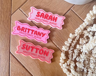 May include: Three pink name tags with white lettering. The name tags are shaped like a banner and have the text "Sarah Registered Nurse", "Brittany Registered Nurse", and "Sutton Registered Nurse".