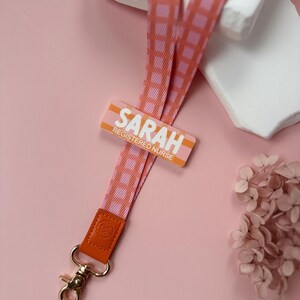May include: A pink and orange lanyard with a repeating checkered pattern. The lanyard has a gold-colored clasp and a leather accent. The name tag reads "SARAH REGISTERED NURSE" in white letters.