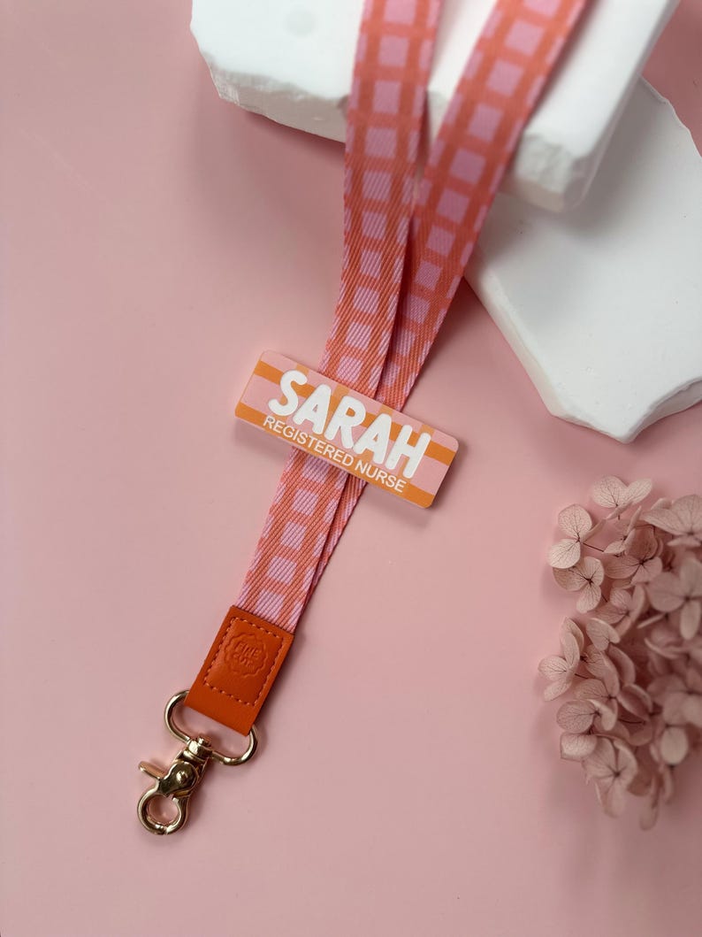 May include: A pink and orange lanyard with a repeating checkered pattern. The lanyard has a gold-colored clasp and a leather accent. The name tag reads "SARAH REGISTERED NURSE" in white letters.