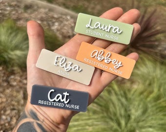 May include: Four rectangular name badges in various colours are displayed. The badges include the names "Laura," "Eliza," "Abbey," and "Cat," along with their respective titles: "Student Nurse," "Anum," and "Registered Nurse." The badges are in shades of green, beige, orange, and grey.