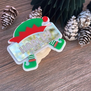 May include: A Christmas-themed name tag in the shape of an elf hat and shoes. The name tag is white with green and red accents, and the name "HARLEY WARD CLERK" is printed on it. The background includes pine cones and a green paper Christmas tree.