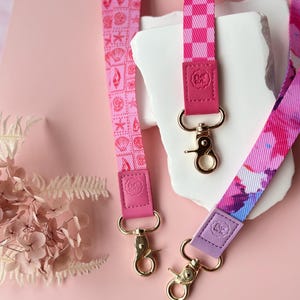 May include: Three pink lanyards with gold-colored clasps. One lanyard features a seashell pattern, another has a checkered design, and the third has a watercolor floral print. Each lanyard has a leather accent with a logo.