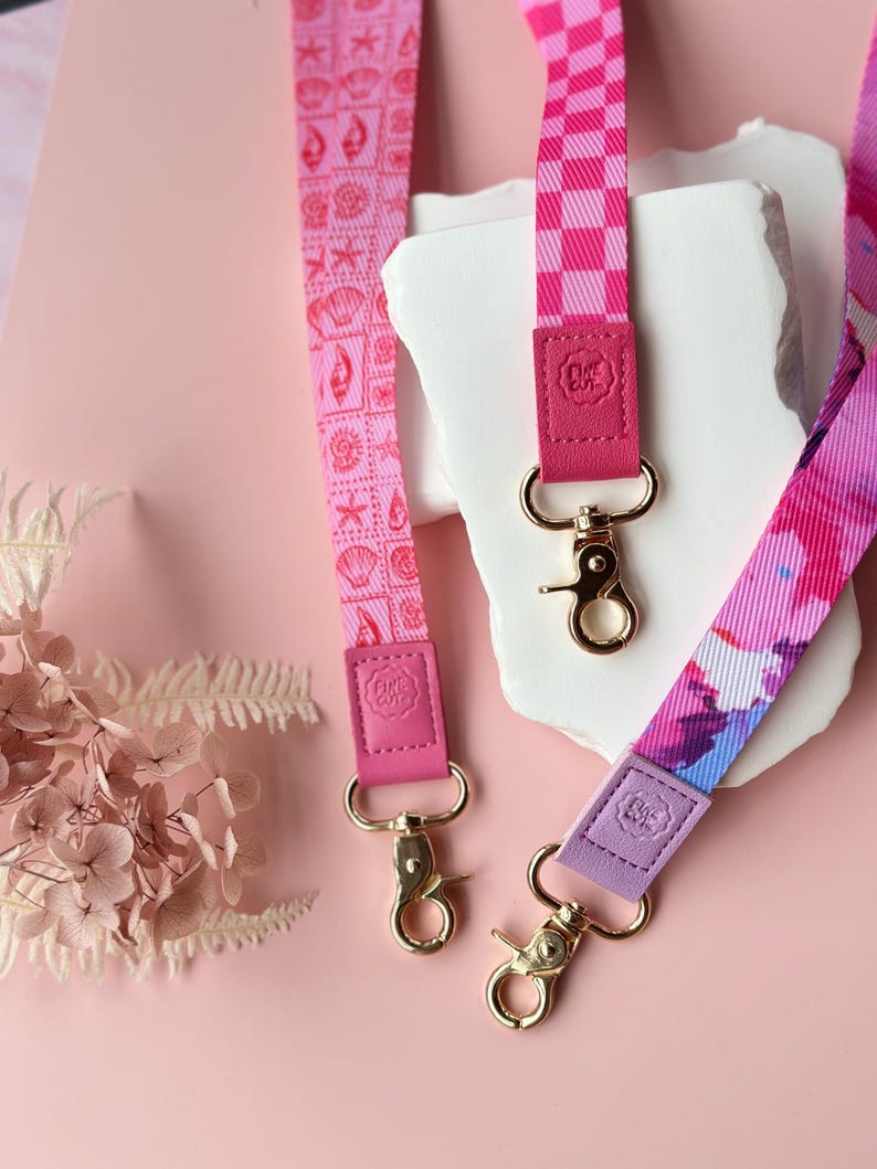 May include: Three pink lanyards with gold-colored clasps. One lanyard features a seashell pattern, another has a checkered design, and the third has a watercolor floral print. Each lanyard has a leather accent with a logo.