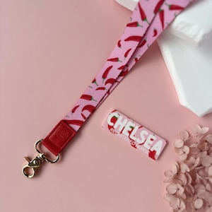 May include: A pink lanyard with a red chili pepper pattern, a red leather accent, and a gold-colored clasp. A matching name tag with the name "CHELSEA" and the title "REGISTERED NURSE" is also present.