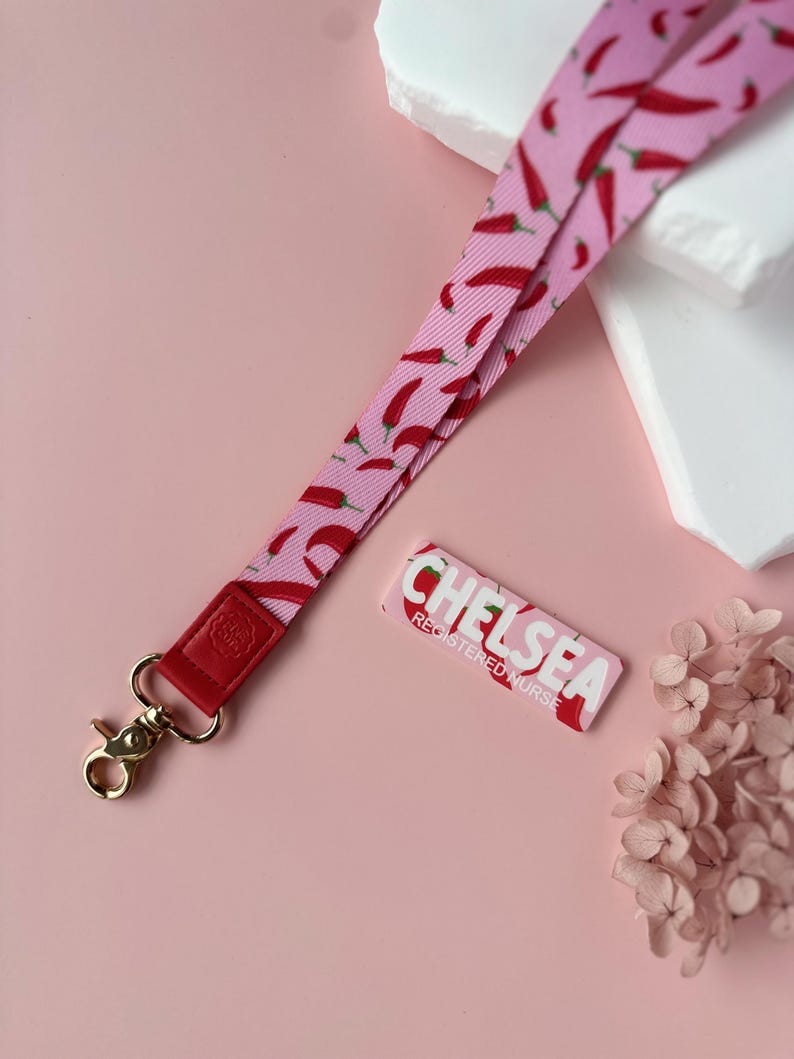 May include: A pink lanyard with a red chili pepper pattern, a red leather accent, and a gold-colored clasp. A matching name tag with the name "CHELSEA" and the title "REGISTERED NURSE" is also present.