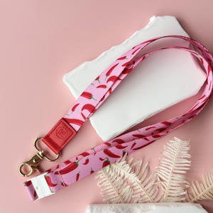 May include: Pink lanyard with a repeating pattern of red chili peppers. The lanyard has a red leather accent with a gold-colored clasp and a white plastic buckle. The background is a light pink color.