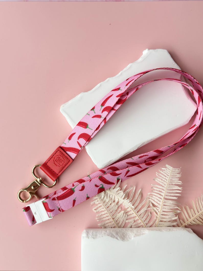 May include: Pink lanyard with a repeating pattern of red chili peppers. The lanyard has a red leather accent with a gold-colored clasp and a white plastic buckle. The background is a light pink color.