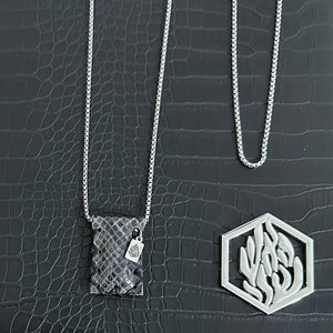 Puede incluir: Un collar de cadena de plata con un colgante de bolsa de piel de serpiente gris y un colgante hexagonal de plata con un diseño floral estilizado.