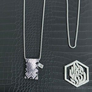 Puede incluir: Dos cadenas de plata con un colgante en una de ellas. El colgante es una pequeña bolsa rectangular con un patrón de piel de serpiente. La otra cadena tiene un colgante hexagonal con un diseño floral estilizado.