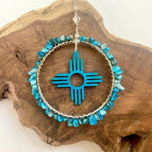May include: A turquoise blue Southwestern-style sun symbol, or "Sun Zia", is surrounded by turquoise blue stones and wire wrapped in a gold-toned metal hoop. The hoop is suspended from a silver chain with a crystal bead.