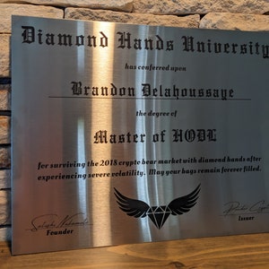 Może przedstawiać: Srebrna metalowa tabliczka z tekstem "Diamond Hands University" i "Master of HODL" w czarnym, staroangielskim kroju pisma. Tabliczka jest certyfikatem przyznanym Brandonowi Delahoussage za przetrwanie kryptowalutowego rynku niedźwiedzia z 2018 roku.