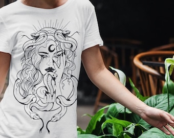 May include: White t-shirt with a black and white illustration of a woman with snakes in her hair. The woman has a crescent moon on her forehead and is holding a snake in her hand.