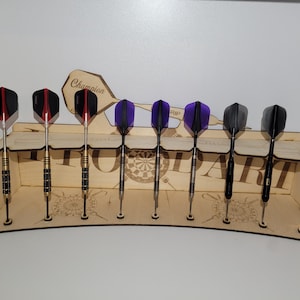 Może przedstawiać: Drewniany uchwyt na rzutki z ośmioma rzutkami. Rzutki mają różne kolorowe lotki, w tym czerwone, czarne, fioletowe i srebrne. Uchwyt ma zakrzywioną konstrukcję z widocznym słowem "DART". Widoczne jest również słowo "Champion".