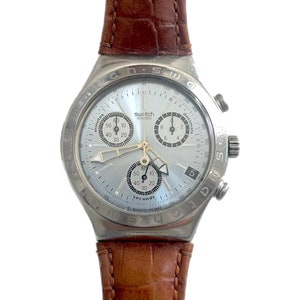 Può includere: Un orologio cronografo Swatch argentato con quadrante chiaro e cinturino in pelle marrone. Il quadrante presenta più quadranti secondari e la scritta "Swatch". Il cinturino è in pelle marrone strutturata.