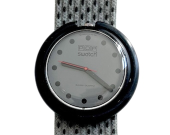 Könnte beinhalten: Eine schwarze Swatch POP Uhr mit einem grauen Zifferblatt und einem grau-weißen gemusterten Armband. Das Zifferblatt der Uhr trägt den Text "POP swatch" und "SWISS QUARTZ".