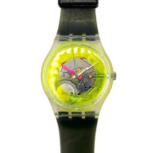 May include: A transparent Swatch watch with a neon yellow face and a black strap. The watch face displays the inner workings of the watch, including gears and a pink second hand. The watch is a classic design.