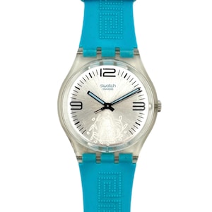 Op de afbeelding: Een Swatch horloge met een zilveren wijzerplaat en een turquoise blauwe siliconen band. De wijzerplaat heeft zwarte cijfers op 12, 3 en 9 uur, met een bloemenmotief. De band heeft een geometrisch patroon.