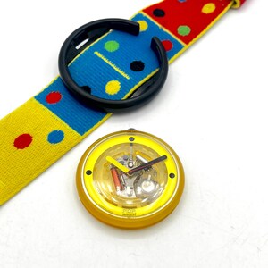 May include: A yellow and black watch with a colorful strap. The watch face is yellow with a clear cover, revealing the internal mechanisms. The strap is patterned with red, blue, and yellow blocks and polka dots. The watch is a POP Swatch.