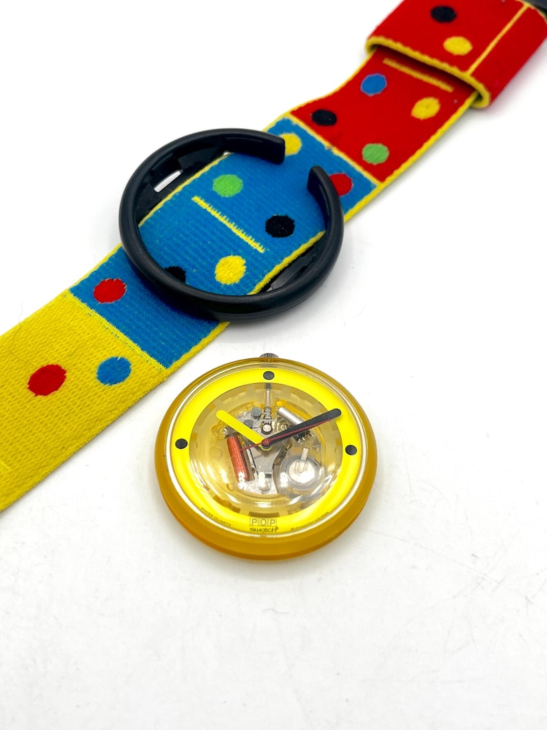 May include: A yellow and black watch with a colorful strap. The watch face is yellow with a clear cover, revealing the internal mechanisms. The strap is patterned with red, blue, and yellow blocks and polka dots. The watch is a POP Swatch.